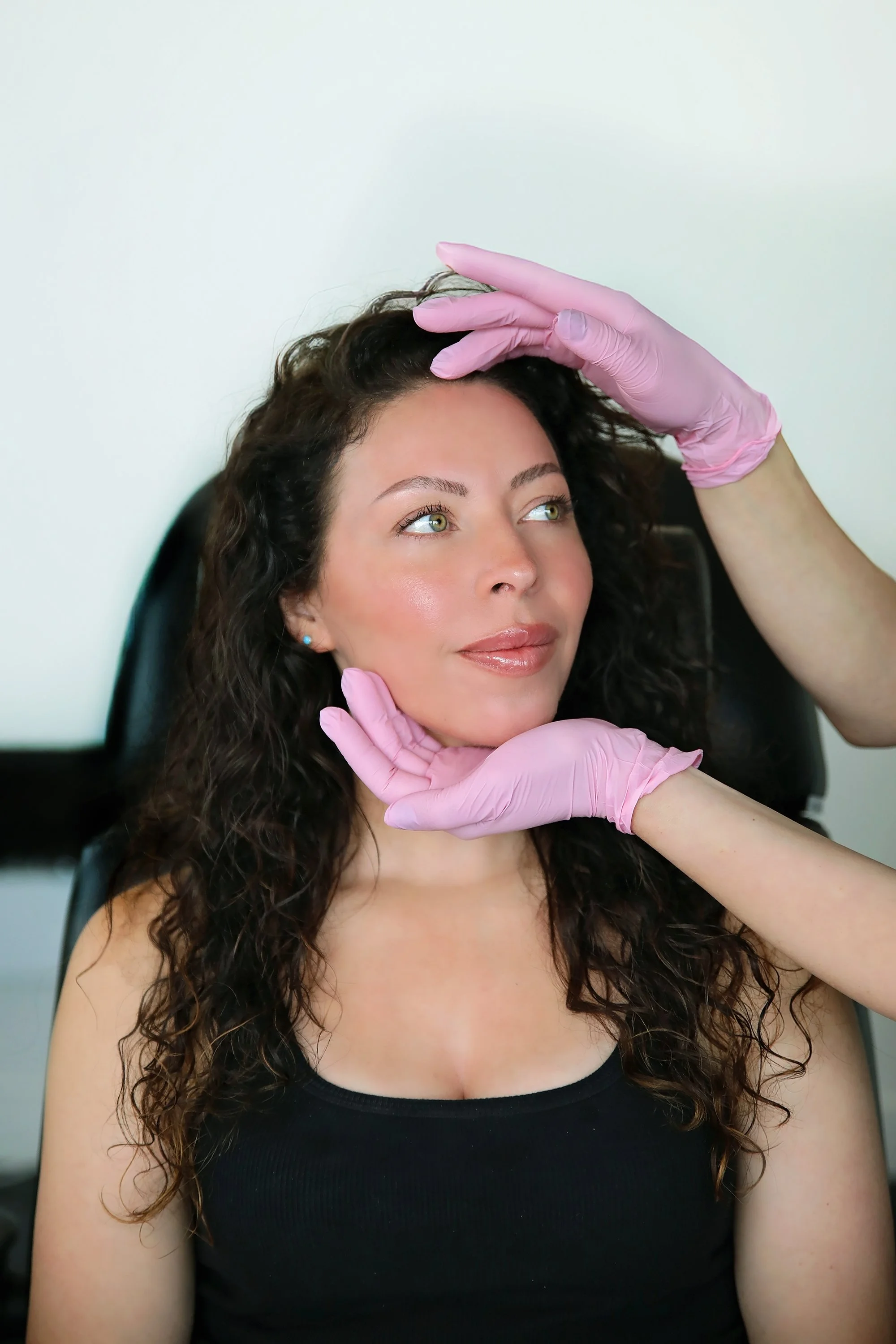 A woman sitting in a chair getting a medical or cosmetic examination. She has curly hair, light skin, and is wearing a black tank top. A person wearing pink gloves is examining her forehead and chin.